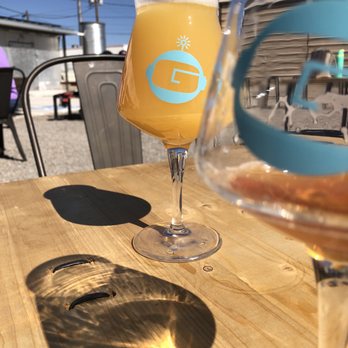 Gravity Bound Brewing - Hazy IPA - A glass of hazy IPA beer with a logo on it is placed on a wooden table outdoors. The glass casts a shadow on the table, and the background includes outdoor chairs, a building, and a clear blue sky. The scene suggests a sunny day at an outdoor seating area of a brewery or bar. - Brewpubs Near Me - Albuquerque, New Mexico