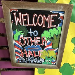 A chalkboard sign with a wooden frame features colorful hand-drawn lettering and illustrations. The sign reads "WELCOME to OTHER HALF BUFFALO" with decorative trees, clouds, and a background of red, white, and blue stripes. It is displayed outdoors or in a casual setting, indicating a welcoming atmosphere for visitors to the brewery.