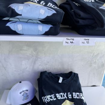 Rice Box & Boba  - Merch - Black and white shirts with Rice Box & Boba logos are displayed on a shelf and folded on a table, with pricing signs for shirts and hats. - Laotian Near Me - Albuquerque, New Mexico