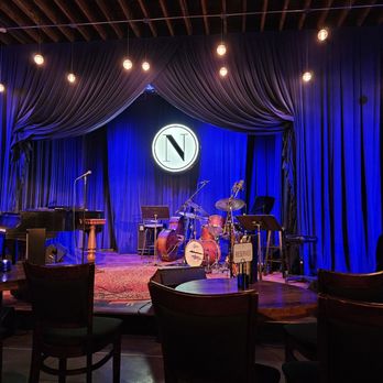 Noce - a stage with a drum set up in front of a clock - Cocktail Bars Near Me - Des Moines, Iowa