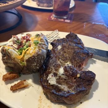 Outback Steakhouse - Ribeye* 13 oz. - A meal featuring a cooked steak, a baked potato topped with cheese, bacon, and green onions, and a glass of soda on a wooden table. - Steakhouses Near Me - Slidell, Louisiana