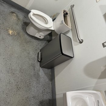 Royyim Thai - Women's Restroom - The image shows a restroom with a white toilet, a small trash can, and a sink. The floor is gray with dirt and debris, including a noticeable spill or stain near the toilet. The toilet paper holder is empty, and the toilet tank lid is missing, exposing internal components. The walls are plain, with metal grab bars installed. The overall condition appears neglected, with signs of dirt and disrepair. - Thai Near Me - Mesa, Arizona