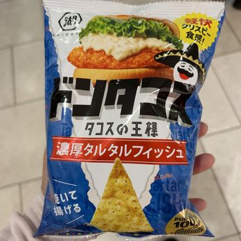 Shibuyala - A snack bag held in a person's hand, displaying Japanese text and graphics. The packaging features an image of a taco with fried fish, lettuce, and sauce, along with a cartoon character wearing a sombrero and smiling. The bag has a blue background with additional Japanese text and a triangular chip illustration at the bottom. The setting appears to be indoors, with tiled flooring visible. - Cosmetics & Beauty Supply Near Me - Arcadia, California
