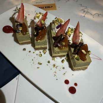 NiMo - Sicilian Pistachio Mousse - Four slices of pistachio mousse cake are arranged on a white rectangular plate. Each slice is decorated with a strawberry wedge, pistachios, edible flowers, and a drizzle of caramel sauce. The plate also features small drops of red sauce and scattered pistachios. A menu or receipt is partially visible in the background on a marble table surface. - Salad Near Me - Tequesta, Florida