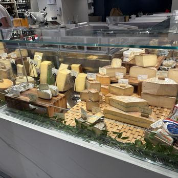 Nouvelle Maison - A glass display case showcases an assortment of cheeses, including hard, soft, and aged varieties, with labels and wooden stands. - Grocery Near Me - Winchester, Massachusetts