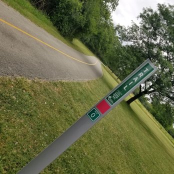 Brookfield Woods - A park sign displaying the word 'PARK' is installed on a metal pole next to a winding paved pathway. The pathway curves through a grassy area with trees and bushes on either side, under an overcast sky. The scene depicts a natural outdoor park environment with lush greenery and a maintained walking trail. - Parks Near Me - Brookfield, Illinois