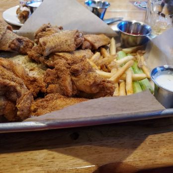 Twin Peaks - Wings - The image shows a metal tray lined with parchment paper containing fried chicken wings, French fries, celery sticks, and small metal cups of dipping sauces. The tray is placed on a wooden table, with additional food items and drinks visible in the background. The setting appears to be a casual dining restaurant, focusing on fried chicken and snack foods. - American Near Me - Albuquerque, New Mexico