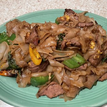 Thai Kitchen - Beef Drunken Noodles - A serving of beef drunken noodles featuring wide rice noodles, cooked with beef slices, green onions, and mixed vegetables such as bell peppers. The dish is presented on a green oval plate, placed on a light-colored textured surface, showcasing traditional Thai stir-fried noodle preparation. - Thai Near Me - Temecula, California