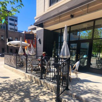 Arbor - Exterior - Arbor restaurant's outdoor patio features tables with chairs and closed umbrellas, enclosed by a black metal fence, with a building entrance and cityscape in the background. - Breakfast & Brunch Near Me - Fort Wayne, Indiana