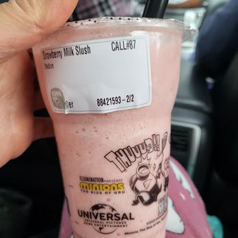 Kung Fu Tea - Strawberry milk slush with cute Minions on the cup - A pink strawberry milk slush in a clear plastic cup with a Minions-themed design, including Minions characters and logos from Universal Pictures. The cup has a label indicating the flavor, size, and a call number, and is held by a person wearing pink clothing with a white pattern. The background shows the interior of a vehicle, with a blurred view of the dashboard and a passenger seat. - Bubble Tea Near Me - Albuquerque, New Mexico