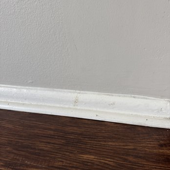 Clean Champs - The image shows a close-up of a white baseboard with some dirt, scuff marks, and slight damage, positioned at the junction of a light gray wall and a dark wooden floor. The baseboard appears to be made of painted wood or MDF, with visible imperfections and signs of wear. The wall has a textured surface, and the floor has a polished wood grain finish. - Home Cleaning Near Me