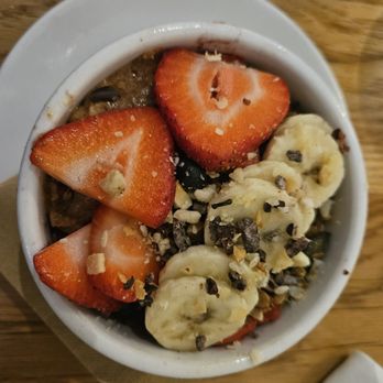SŌW Plated - Acai bowls - A bowl of oatmeal topped with sliced strawberries, banana slices, chopped nuts, and seeds, served on a wooden table. - New American Near Me - Upper Arlington, Ohio
