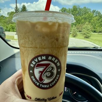 7 Brew Coffee - A plastic cup with iced coffee from Seven Brew Drive Thru Coffee, featuring the logo and a red straw, is held inside a car. - Coffee & Tea Near Me - Brownsburg, Indiana