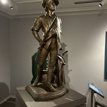 Concord Museum - The image shows a bronze statue of a young girl dressed in pioneer or explorer clothing, including a wide-brimmed hat, shirt, and pants, holding a rifle in her right hand. The statue is positioned on a raised platform in a museum setting, with a gray wall and informational signage visible nearby. The sculpture captures a confident stance, with her left hand resting on her hip and a bag or bundle at her feet. The lighting highlights the detailed features and clothing of the statue. - Museums Near Me - Concord, Massachusetts
