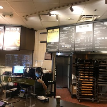 Panera Bread - Dinner - A Panera Bread cashier, wearing a face mask and gloves, is positioned behind the counter with a coffee cup in front. The background shows menu boards and bakery racks. - Sandwiches Near Me - Leesburg, Virginia
