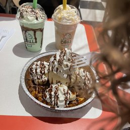 Grasshopper - The image shows two milkshakes in clear cups with dome lids and colorful straws, one green and one yellow, on a table at Chill Milkshake Bar. In front, there is a dessert in a foil container topped with whipped cream, banana slices, and chocolate syrup, with a fork inserted into it. The background includes a person and a checkered floor pattern.