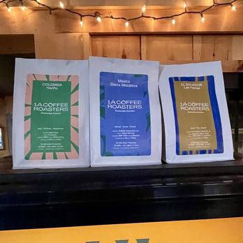 1a Coffee Roasters - The beans - Three white paper bags of coffee beans are placed on a black counter, each with a distinct colorful label indicating their region of origin: Colombia, Mexico, and El Salvador. The labels include details about the coffee's origin and the roastery. The background features a wooden wall with string lights hanging above, creating a warm ambiance. - Coffee & Tea Near Me - West Dover, Vermont