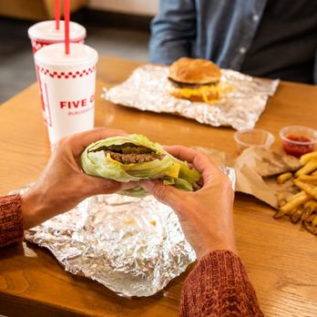 Five Guys - A customer's hands hold a Five Guys lettuce wrap at a dining room table inside a Five Guys restaurant. - Burgers Near Me - Red Deer, Alberta Five Guys - A customer's hands hold a Five Guys lettuce wrap at a dining room table inside a Five Guys restaurant. - Burgers Near Me - Red Deer, Alberta