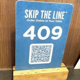 The option to order online instead of waiting in line. - A blue sign with the text 'Skip the Line' and 'Order Online at Your Table,' displaying table number 409 and a QR code, placed on a wooden stand.