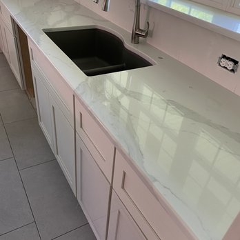 Montes Marble & Granite - Kitchen sink quartz countertop - A kitchen countertop made of white marble-look material with gray veining, featuring a black undermount sink and a stainless steel gooseneck faucet. The countertop is installed beneath a window that provides natural light and a view of green foliage outside. Electrical outlets are visible on the wall above the counter, and the cabinetry below is painted white with a paneled design. The floor consists of large gray tiles. - Building Supplies Near Me - Atkinson, New Hampshire
