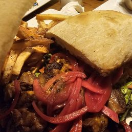 Braised Short Rib - A close-up of a sandwich with toasted bread, accompanied by French fries and topped with sliced red bell peppers and vegetables. The plate is on a wooden table with other dishes and condiments visible in the background, indicating a restaurant setting.