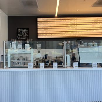 Carte Blanche - Interior - The image shows a cafe counter with a white, vertically ridged front panel. Behind the counter, there is coffee brewing equipment and supplies visible through a glass barrier. A menu board with various coffee and beverage options is mounted on the wall above, with prices listed. The ceiling has recessed lighting and a decorative strip of light. The overall setting appears clean and modern, with a focus on coffee service. - Coffee & Tea Near Me - Mountain View, California