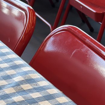 Mighty Fine Burgers Cedar Park - Chairs - The image shows multiple red metal chairs with shiny, reflective surfaces arranged around a table. The table is covered with a blue and white checkered fabric tablecloth. The chairs are stacked or placed closely together, and the setting appears to be indoors, possibly in a restaurant or cafe. - Burgers Near Me - Cedar Park, Texas