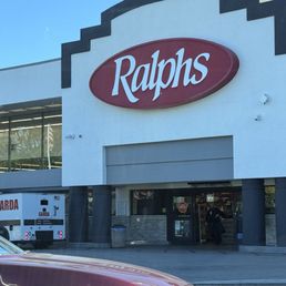 The photo captures the storefront of a Ralphs grocery store, featuring a large red and white sign with the Ralphs logo. The building has a white facade with black accents and a partially visible entrance with automatic doors. A vehicle is parked in front, and a Garda security vehicle is also visible nearby. The sky is clear and blue, indicating a sunny day.