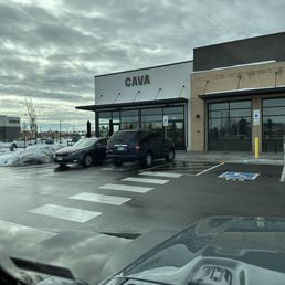 Photo shows the front of CAVA, a Mediterranean-inspired restaurant, with a parking lot in the foreground. Several cars are parked, including a black SUV and a sedan. There is a designated handicap parking space visible. The building has large glass windows and a sign displaying the name 'CAVA'. The sky is overcast with gray clouds, and some snow is present on the ground and around the parking area.