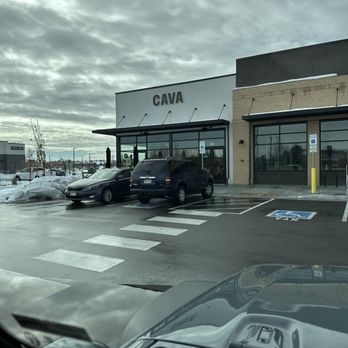 CAVA - Photo shows the front of CAVA, a Mediterranean-inspired restaurant, with a parking lot in the foreground. Several cars are parked, including a black SUV and a sedan. There is a designated handicap parking space visible. The building has large glass windows and a sign displaying the name 'CAVA'. The sky is overcast with gray clouds, and some snow is present on the ground and around the parking area. - Mediterranean Near Me - Denver, Colorado