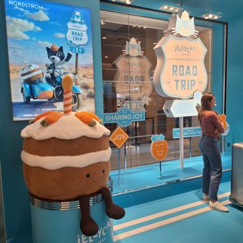 Jellycat - A plush cupcake character with a smiling face, striped candle, and brown legs is placed on a cylindrical stand with the Jellycat logo. Behind it, a store window displays the Jellycat Road Trip theme with signage and a large poster featuring a cartoon cat riding a motorcycle with a sidecar. A woman stands nearby, looking at the window display. The setting has a bright, modern interior with blue walls and ceiling accents. - Toy Stores Near Me - Chicago, Illinois