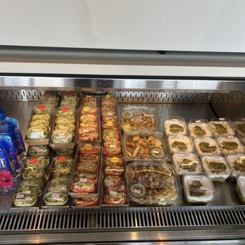 Dar Coffee - Fresh to go food - The image shows a refrigerated display case containing various pre-packaged salads, wraps, and prepared foods. The items are organized in clear plastic containers and trays, with some labels visible. The display is part of a grocery or deli section, with a metal and wood design. The products include salads with greens, vegetables, and proteins, as well as other ready-to-eat items. - Coffee & Tea Near Me - Plano, Texas