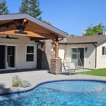 Vimahemo Construction - Construction complete. - The image shows a backyard with a swimming pool surrounded by a concrete patio. There is a house with a covered porch supported by wooden beams, with sliding glass doors leading inside. The yard includes a grassy area, trees, and outdoor furniture, with a clear blue sky overhead. - General Contractors Near Me - Parlier, California
