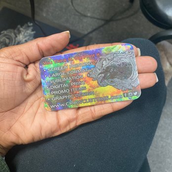 Crow Customs - A hand holds a holographic business card for Crow Customs, featuring printing services like screen printing, sublimation, and graphic design. - Printing Services Near Me - Palmdale, California