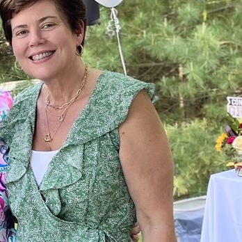 Covet - A woman with short brown hair, wearing a green patterned dress with ruffles and a waist tie, is smiling outdoors at a celebration. Behind her, there are balloons, including one with the word "MAD" and a white balloon, as well as a table with a floral arrangement. The setting appears to be a garden or park with trees and grass. - Women's Clothing Near Me - Boston, Massachusetts