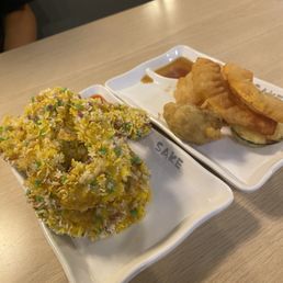 Calimari and veggie tempura - The photo features two white dishes on a light wooden table. One dish contains fried vegetable tempura, including zucchini and other vegetables, with a small container of dipping sauce. The other dish holds a large, crispy fried rice ball decorated with multicolored sprinkles, likely sweet or savory. The setting appears to be a casual dining restaurant, with a blurred background and part of a person's arm visible.