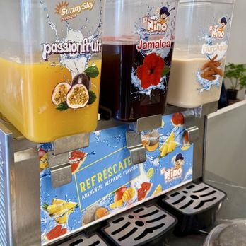 Chicka Loca - Drinks - Three beverage dispensers are positioned on a counter, each containing different drinks: passionfruit, Jamaica (hibiscus), and horchata. The dispensers have colorful labels with images of fruits and ingredients, and the drinks inside vary in color from yellow to dark red and beige. The dispensers are part of a setup promoting authentic Hispanic flavors, with a decorative sign below them. The background includes a plant and a gray wall, indicating an indoor setting. - Peruvian Near Me - Fairfax, Virginia