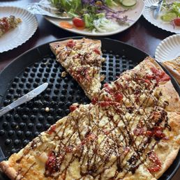 Tuscan pizza, tossed salad - A cheese pizza with toppings and drizzle is on a black tray, surrounded by salad and plates with food on a dining table.