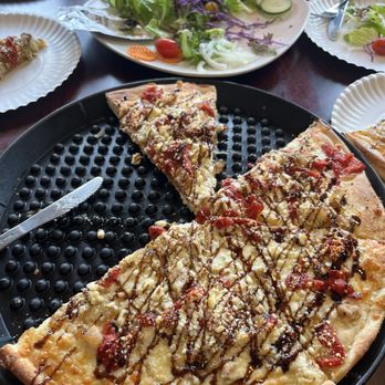 Antigoni's - Tuscan pizza, tossed salad - A cheese pizza with toppings and drizzle is on a black tray, surrounded by salad and plates with food on a dining table. - Pizza Near Me - Lewiston, Maine