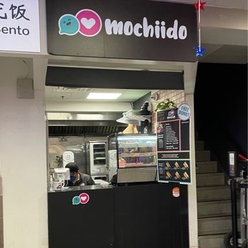 Mochiido - Mochiido is a small dessert shop located inside a food hall, with a black counter and a menu display board. The shop offers Asian fusion bakery treats such as mochi donuts, macarons, and unique items like Ube Tres Leches cake. The signage includes Sanrio-themed desserts like Hello Kitty Strawberry Shortcake and Pompompurin Banana Pudding. A staff member wearing a mask is behind the counter, and a staircase is visible to the right. The shop has limited seating and seasonal designs, with a small American flag and star decorations hanging above. - Donuts Near Me - Flushing, New York
