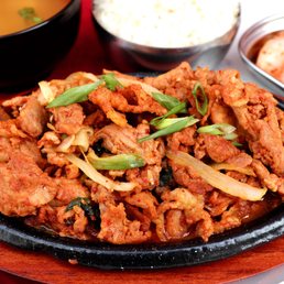 Spicy Pork - A hot, black sizzling plate contains spicy pork with onions and green onions, accompanied by rice and side dishes like kimchi and soup.