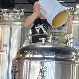 Buckets o' Honey - A person is pouring a liquid from a white bucket into a brewing vessel in a brewery setting, with large stainless steel tanks in the background.