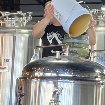 Mad Hatchet Brewing - Buckets o' Honey - A person is pouring a liquid from a white bucket into a brewing vessel in a brewery setting, with large stainless steel tanks in the background. - Breweries Near Me - Shorewood, Illinois