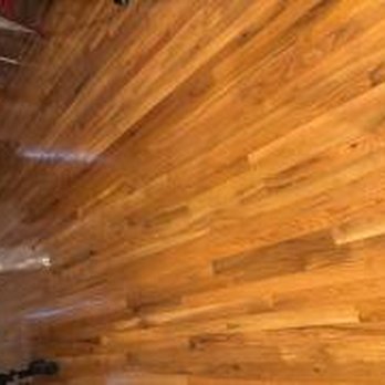 DREYCO Flooring & Design - Oak installed in a large part of the home, replacing carpet. - The image shows a section of a residential interior with a polished hardwood floor featuring a warm, natural wood finish. The floorboards are arranged in parallel, showcasing the wood grain and texture. In the background, there are some furniture pieces and household objects, indicating a living space or similar room. The lighting highlights the smooth surface and rich color of the wood. - General Contractors Near Me - Eugene, Oregon