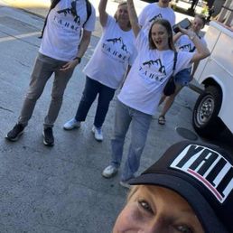 Five individuals wearing matching T-shirts with a mountain logo and text are outdoors, with some making gestures and smiling, while a woman in the foreground takes a selfie.