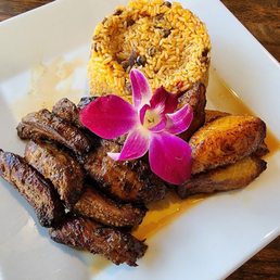 Churrasco - A dish featuring grilled meat, a mound of rice with beans, and fried plantains, garnished with a pink orchid flower, served on a white plate.