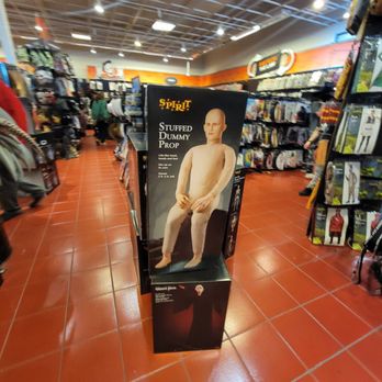 Halloween Express - A life-sized stuffed dummy prop with a human-like appearance, bald head, and neutral facial expression is displayed on a stand in a retail store aisle. The store has a high ceiling with exposed beams and bright lighting. Shelves on both sides contain various products, and shoppers are visible in the background. The dummy is part of the store's Halloween or costume section. - Costumes Near Me - Normal, Illinois