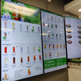 Austea - Menu - Three digital menu displays are mounted on a wall inside Austea in Austin, TX. The screens list a variety of beverages including milk teas, fruit teas, smoothies, and coffee, along with food options like fried chicken wings and sides. The menus include prices, item descriptions, and customization options. The background includes a wall with decorative patterns and a planter with greenery. The setup suggests a modern, self-service ordering environment. - Bubble Tea Near Me - Austin, Texas