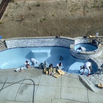 Cerda Pool Construction - During plaster - Multiple construction workers are working on installing a swimming pool and a hot tub in a backyard. The pool has a curved shape with a built-in step, and the hot tub is elevated with a stone exterior. Tools, buckets, and construction materials are scattered around the area, and the surrounding yard is unfinished with dirt and a fence. - Pool & Hot Tub Service Near Me