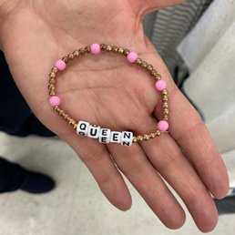 A beaded bracelet featuring pink, gold, and white beads spelling out "QUEEN" is displayed in a person's hand. The bracelet is composed of small round beads, with larger pink beads interspersed among gold beads, and white letter beads forming the word. The person holding the bracelet is wearing a watch on their wrist. The background appears to be an indoor setting, possibly a store or public space, with blurred elements and a tiled floor.